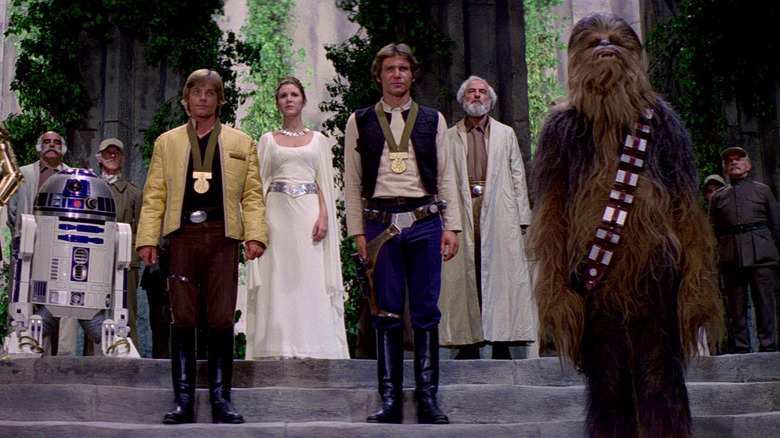Luke Skywalker, vêtu d'une veste jaune, et Han Solo, vêtu d'un gilet bleu, portent des médailles d'or alors qu'ils se tiennent devant la princesse Leia et les autres forces rebelles. 