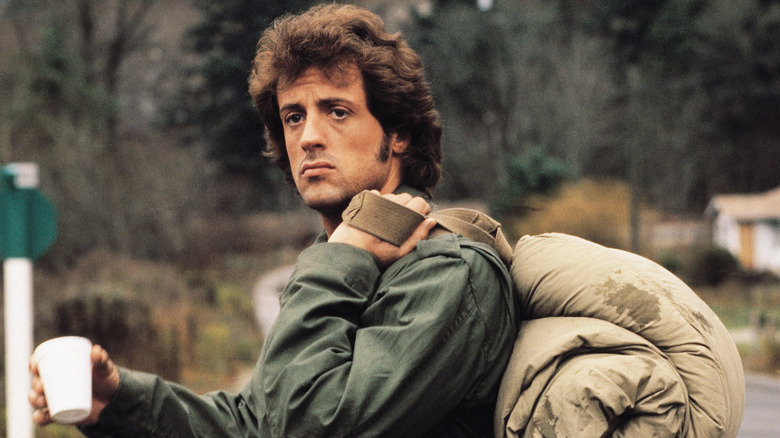 Rambo debout au bord de la route avec une tasse de café et un sac sur le dos dans First Blood (1982)