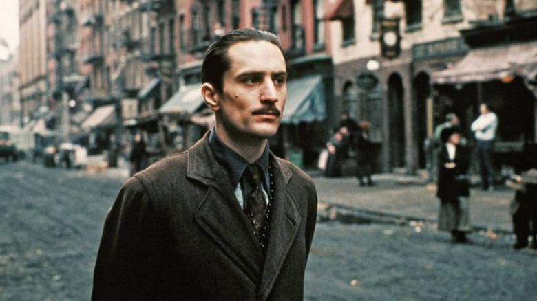 Un jeune Vito Corleone se promène dans les rues de New York des années 1900 dans une veste marron. 