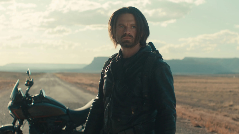 Bucky Barnes se tient sur une route au milieu du désert dans une veste noire devant une moto 