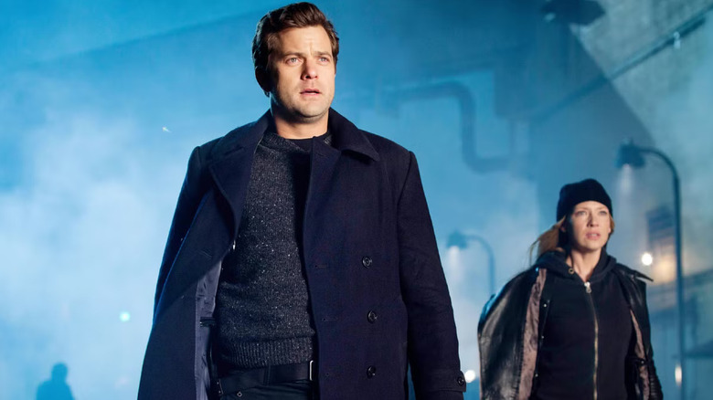 Peter Bishop porte une veste et un pull bleus dans une rue brumeuse 