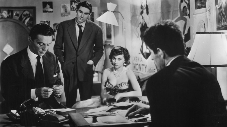 Deux hommes en costume regardent un troisième homme, tandis qu'une femme en robe est assise à proximité, dans 