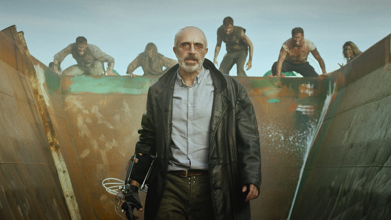 Un homme en trench-coat se tient dans un conteneur métallique tandis que d'autres personnes avancent derrière lui. 