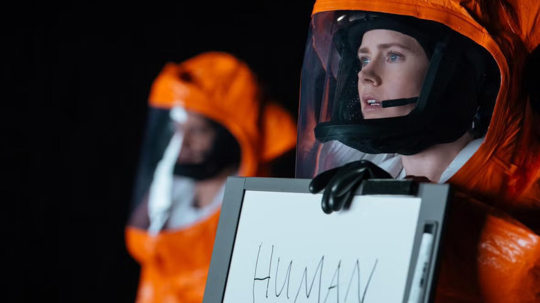 Louise en costume tendant un tableau blanc aux extraterrestres à l'arrivée