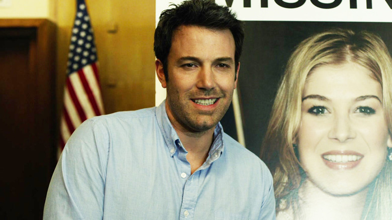 Nick Dunne souriant devant une affiche manquante sur Gone Girl
