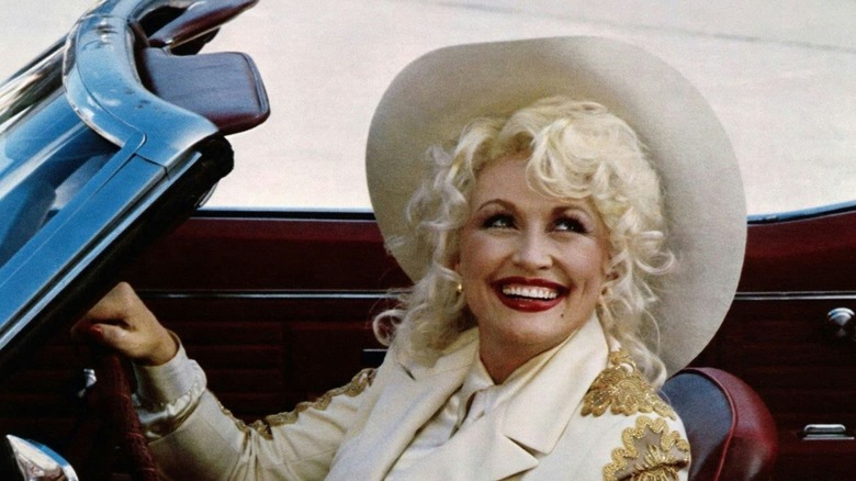 Mona conduisant une voiture et portant un chapeau souriant dans Le meilleur petit bordel du Texas