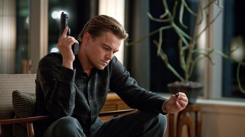 Don Cobb tenant un pistolet et une simple toupie, assis sur une chaise, dans Inception (2010)